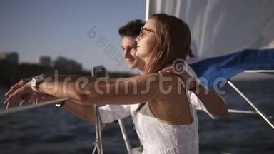 一对年轻夫妇在游艇上共度时光的侧视图。一个穿着白裙子戴着太阳镜坐在一起的女孩
